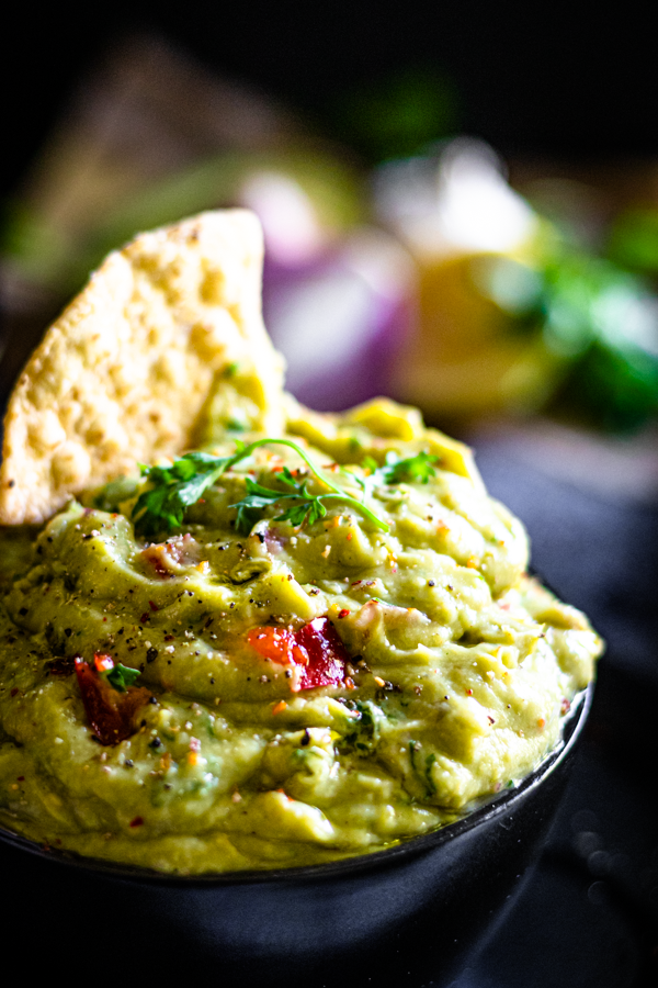 Close-up of a bowl of homemade guacamole with a tortilla chip dipped inside, surrounded by fresh ingredients — ripe avocados, red onion, and lime. A creamy, wholesome guacamole made with cilantro and chilli.