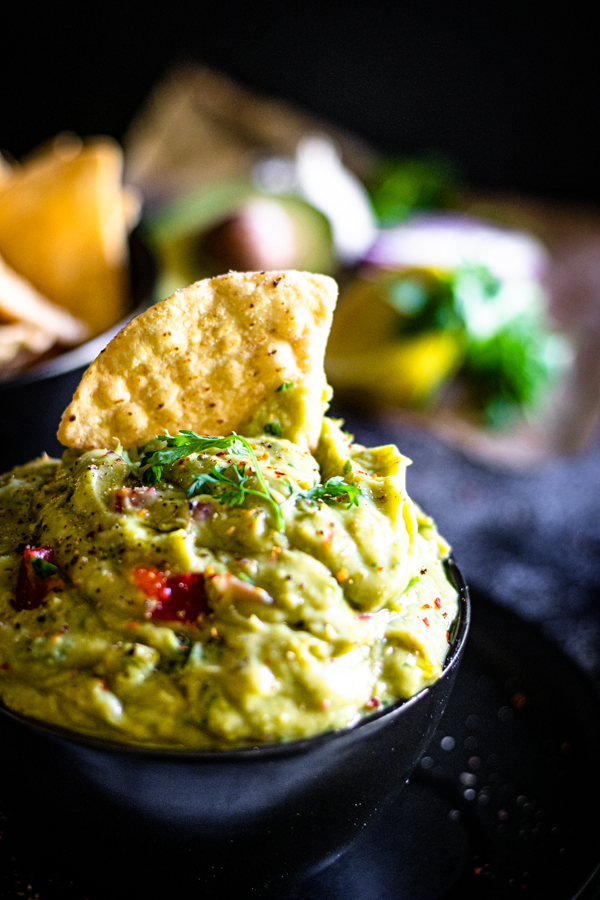 Close-up of creamy guacamole topped with cilantro, a tortilla chip dipped into the bowl, and fresh avocados, red onion, and lime in the background.