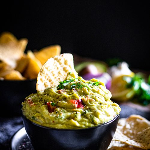 Bowl of homemade guacamole surrounded by tacos, lime, avocado, and red onion — a wholesome and easy guacamole recipe made with ripe avocados, coriander and chilli.