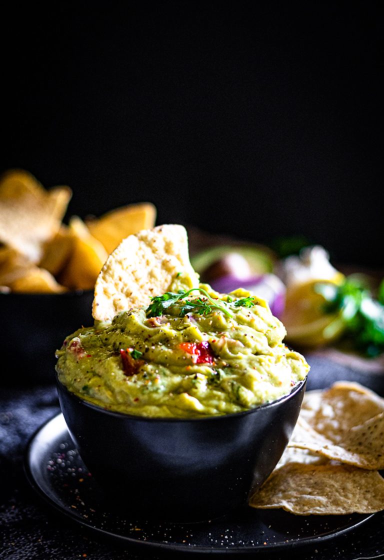 Bowl of homemade guacamole surrounded by tacos, lime, avocado, and red onion — a wholesome and easy guacamole recipe made with ripe avocados, coriander and chilli.