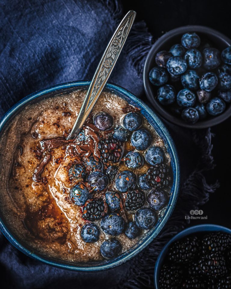 Fermented sorghum porridge (ting ya mabele) served warm in a bowl, topped with blueberries, blackberries, dark chocolate and maple syrup drizzle.
