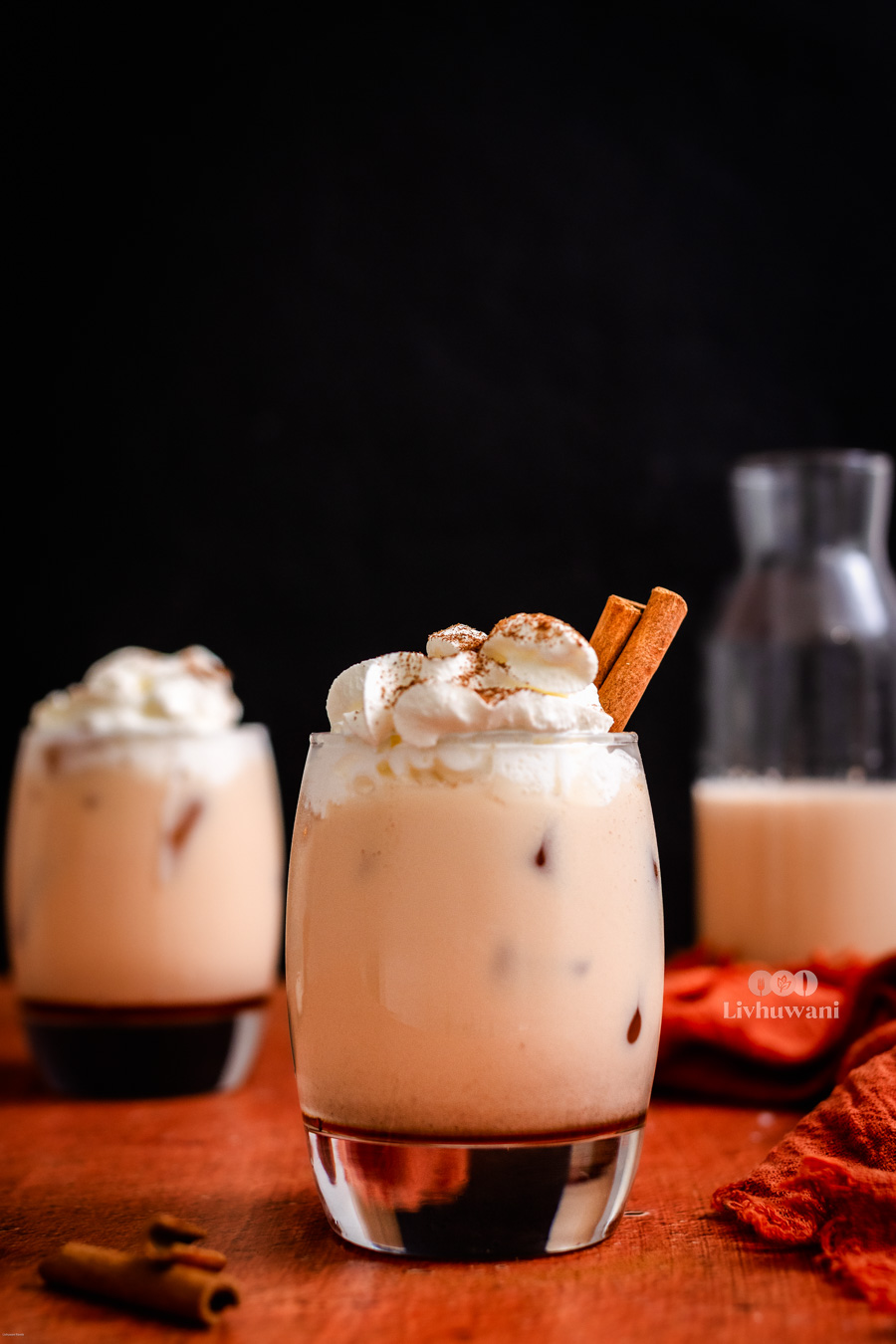Iced rooibos tea latte with whipped cream and cinnamon sticks, served in a glass on a wooden table. Another similar drink and a carafe in the background, ideal for a caffeine-free herbal beverage choice.