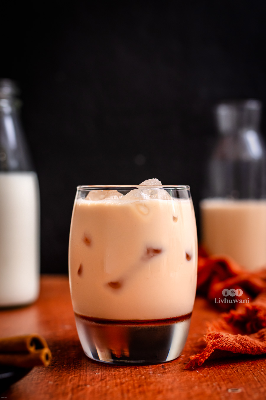 Ice-filled glass of creamy Rooibos tea latte on a wooden table with milk bottles in the background, showcasing a caffeine-free herbal drink option.