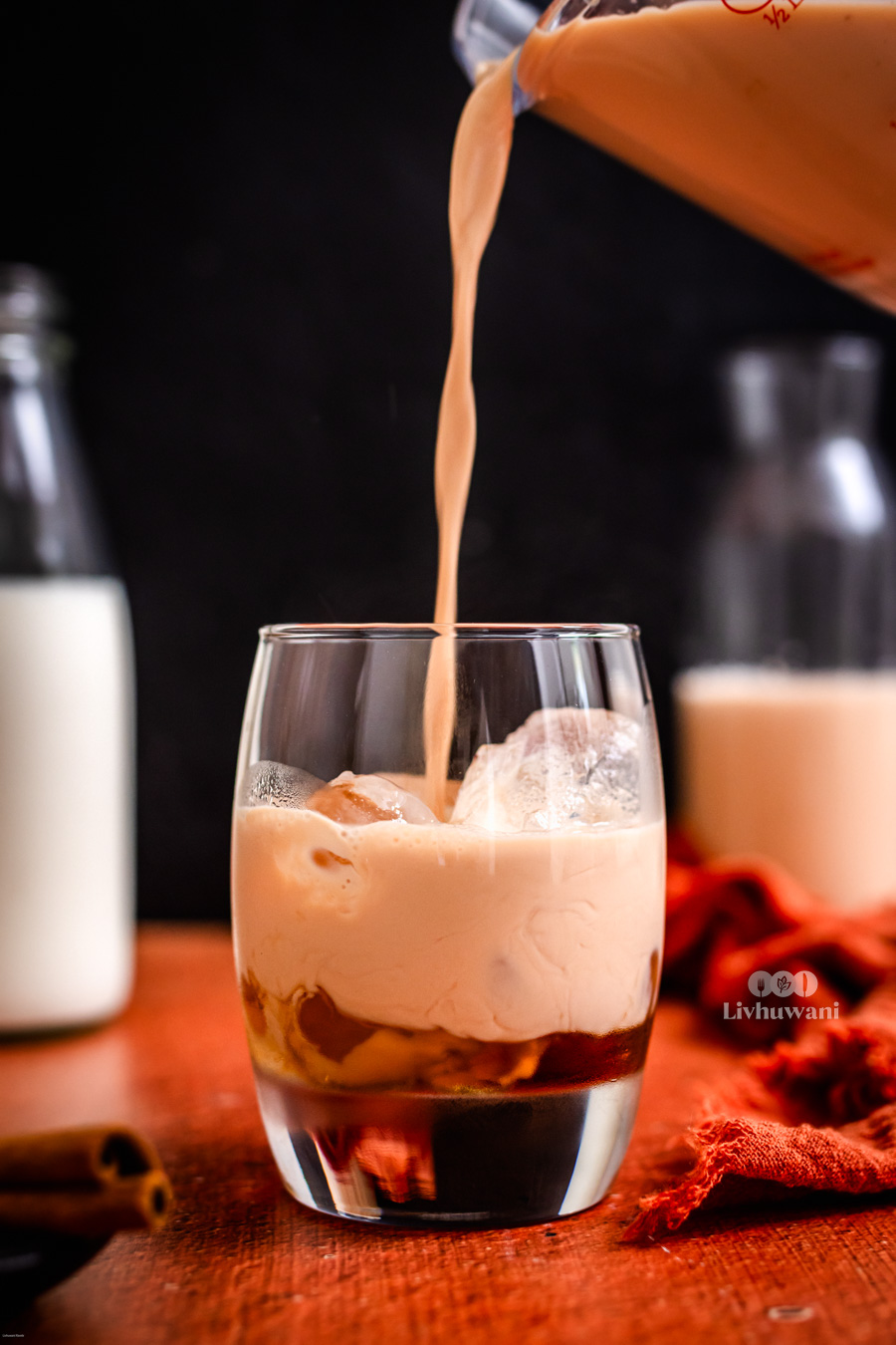 A creamy rooibos tea latte being poured into a glass filled with ice, set on a wooden table. In the background, there are milk bottles and a red cloth, enhancing the warm and inviting aesthetic of this herbal, caffeine-free drink recipe.